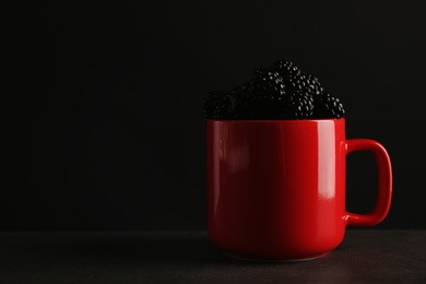 Photo of Mug with fresh blackberry on gray table against dark background. Space for text