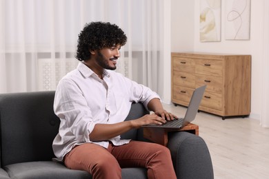 Photo of Happy man using laptop on sofa with wooden armrest table at home