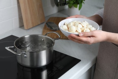 Photo of Woman putting frozen dumplings into saucepan with boiling water on cooktop in kitchen, closeup