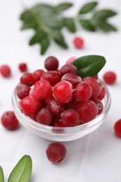 Frozen red cranberries in bowl and green leaves on white table, closeup