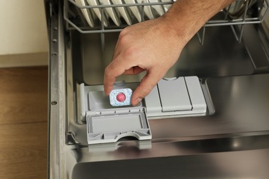 Photo of Man putting detergent tablet into open dishwasher indoors, closeup