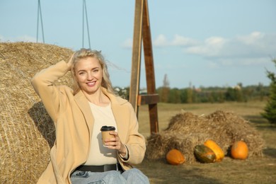 Photo of Beautiful woman with cup of hot drink sitting near hay bale outdoors, space for text. Autumn season