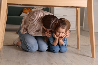 Photo of Scared mother with her little daughter hiding under table in living room during earthquake