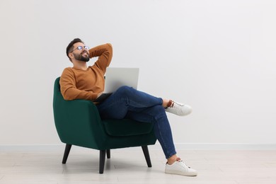 Photo of Handsome man with laptop relaxing in armchair near white wall indoors, space for text