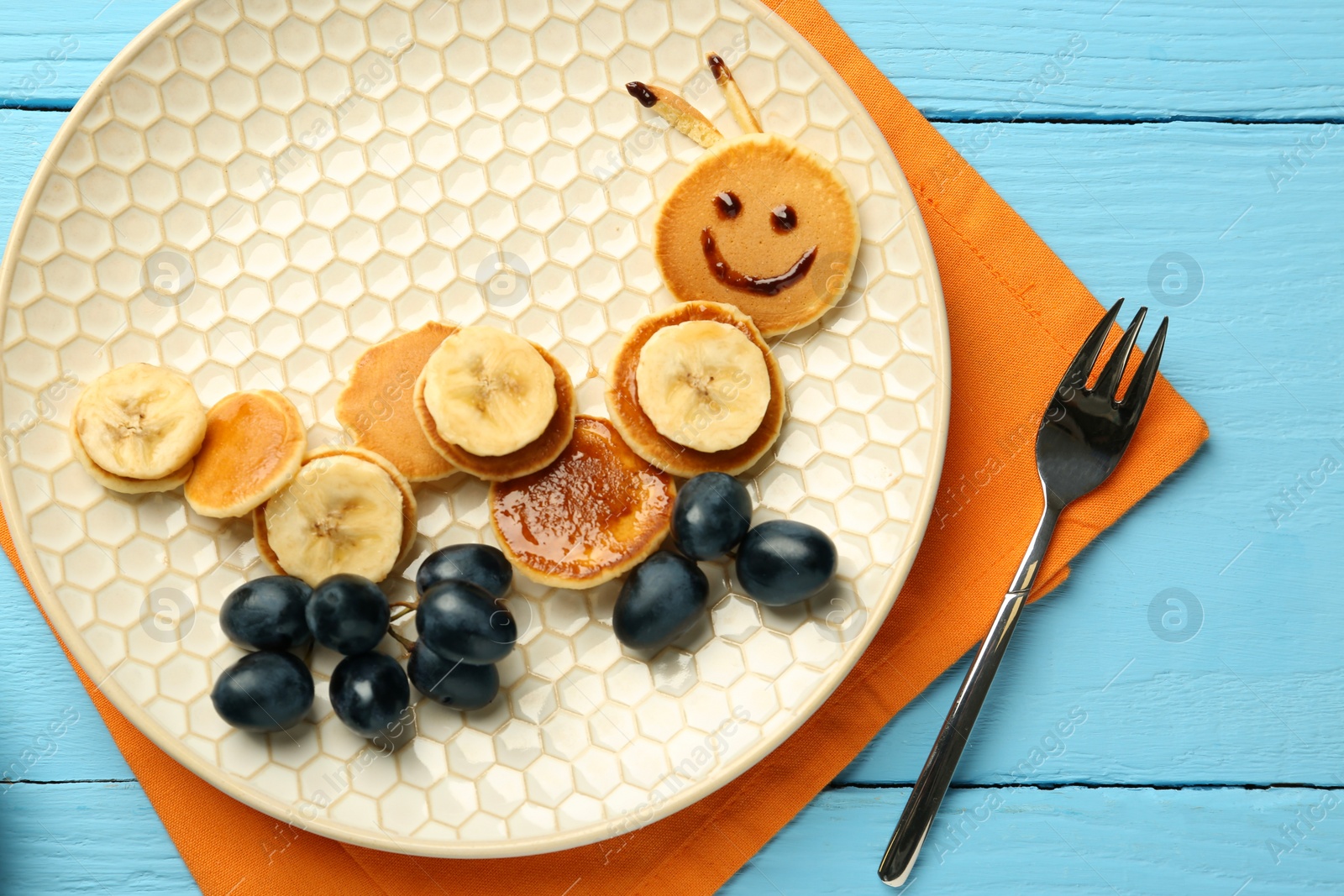 Photo of Creative serving for kids. Plate with cute caterpillar made of pancakes, grapes and banana on light blue wooden table, top view