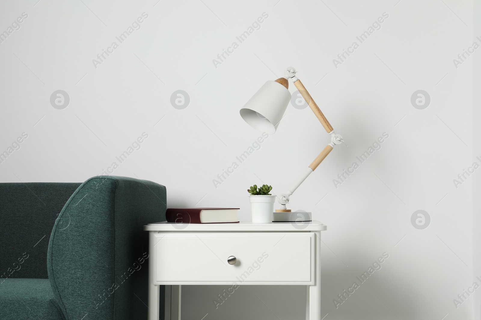 Photo of Stylish lamp, houseplant with book on side table and soft sofa near white wall. Interior design