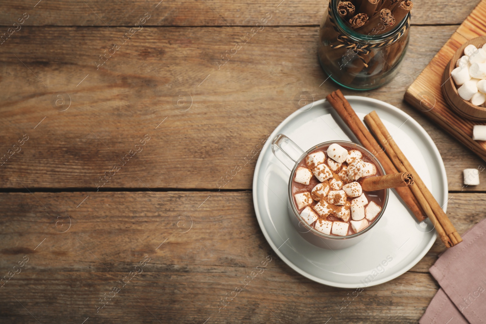 Photo of Glass cup of hot cocoa with aromatic cinnamon and marshmallows on wooden table, flat lay. Space for text