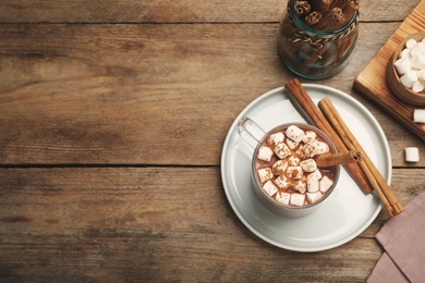 Photo of Glass cup of hot cocoa with aromatic cinnamon and marshmallows on wooden table, flat lay. Space for text