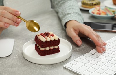 Photo of Bad habits. Woman eating piece of cake while working on computer at light grey table, closeup