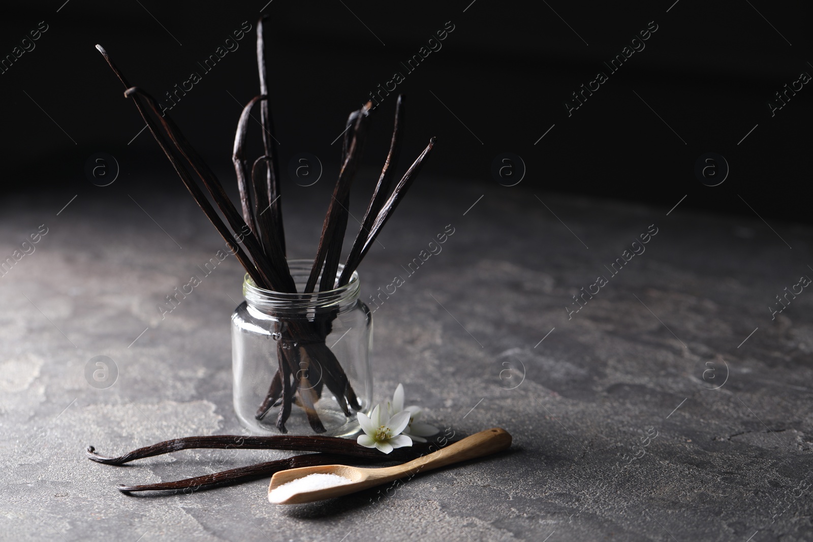 Photo of Spoon with sugar, flowers and vanilla pods on grey textured table, closeup. Space for text