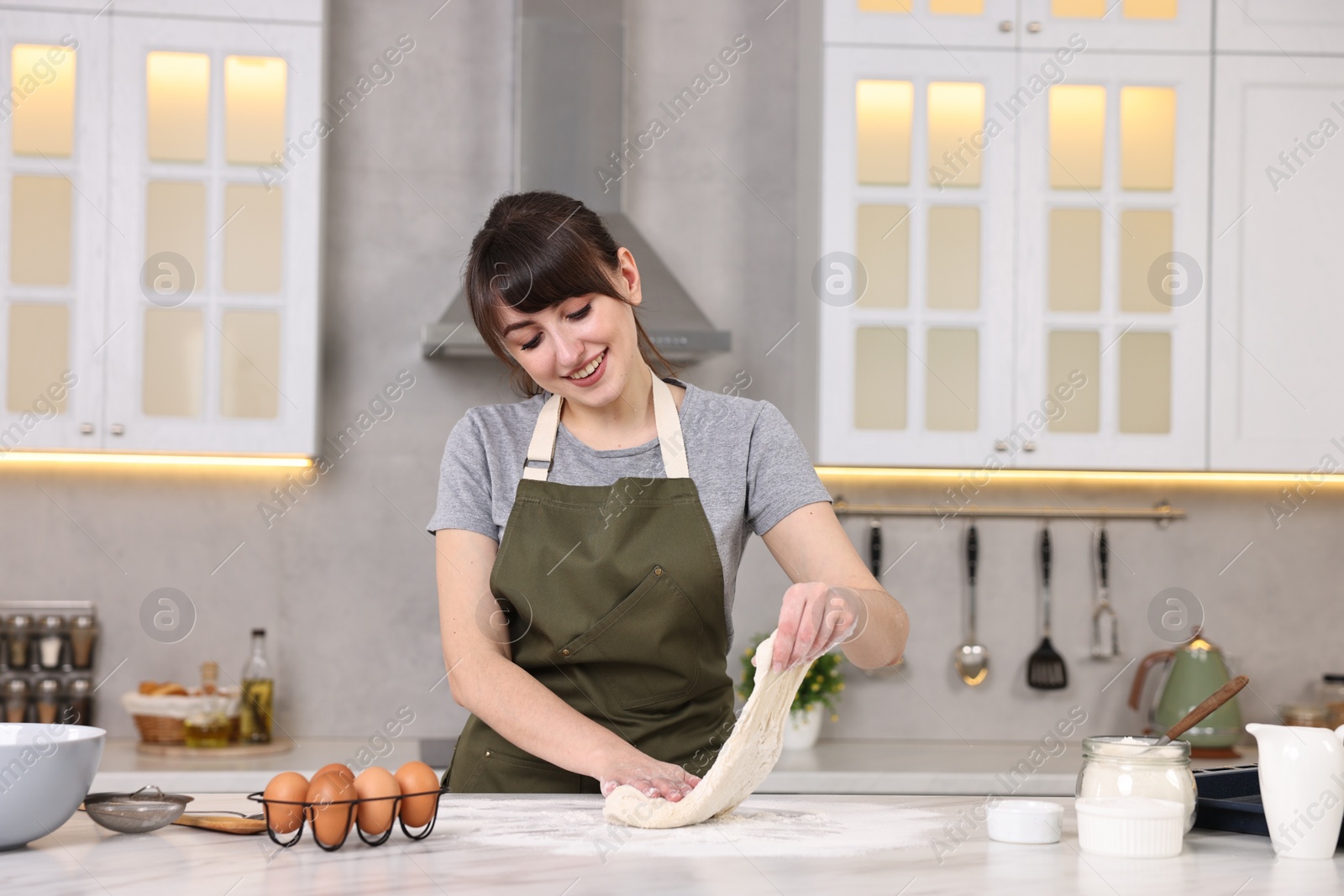 Photo of Happy young housewife kneading dough at white marble table in kitchen