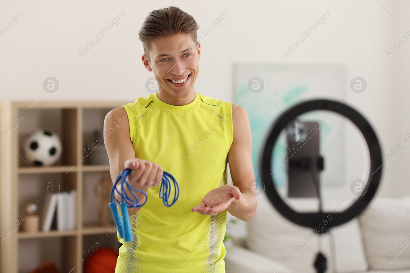 Photo of Smiling sports blogger holding skipping rope while streaming online fitness lesson with smartphone at home