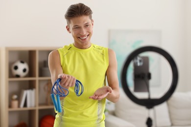 Smiling sports blogger holding skipping rope while streaming online fitness lesson with smartphone at home