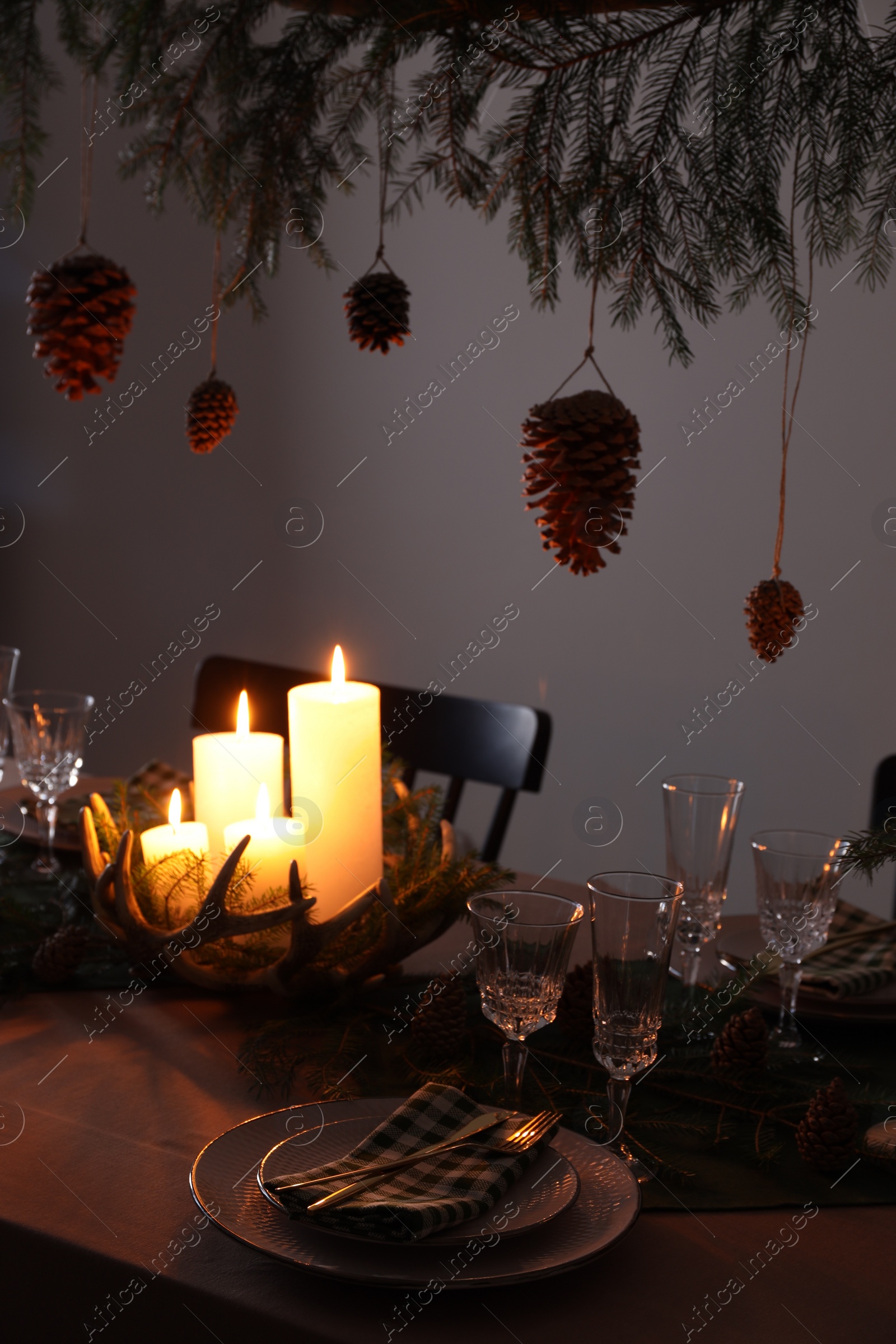 Photo of Christmas celebration. Cones hanging from fir tree branch over table with burning candles and tableware