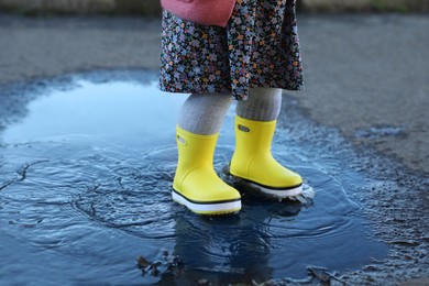 Photo of Little girl wearing rubber boots walking in puddle outdoors, closeup