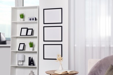 Photo of Stylish room interior with empty frames hanging on white wall near shelving unit