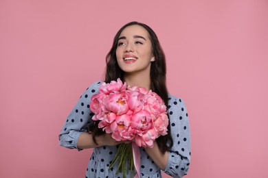 Beautiful young woman with bouquet of peonies on pink background