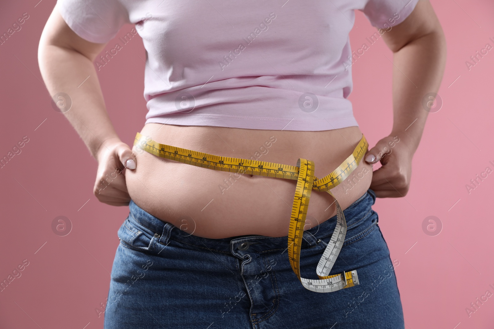 Photo of Woman with measuring tape touching belly fat on pink background, closeup. Overweight problem