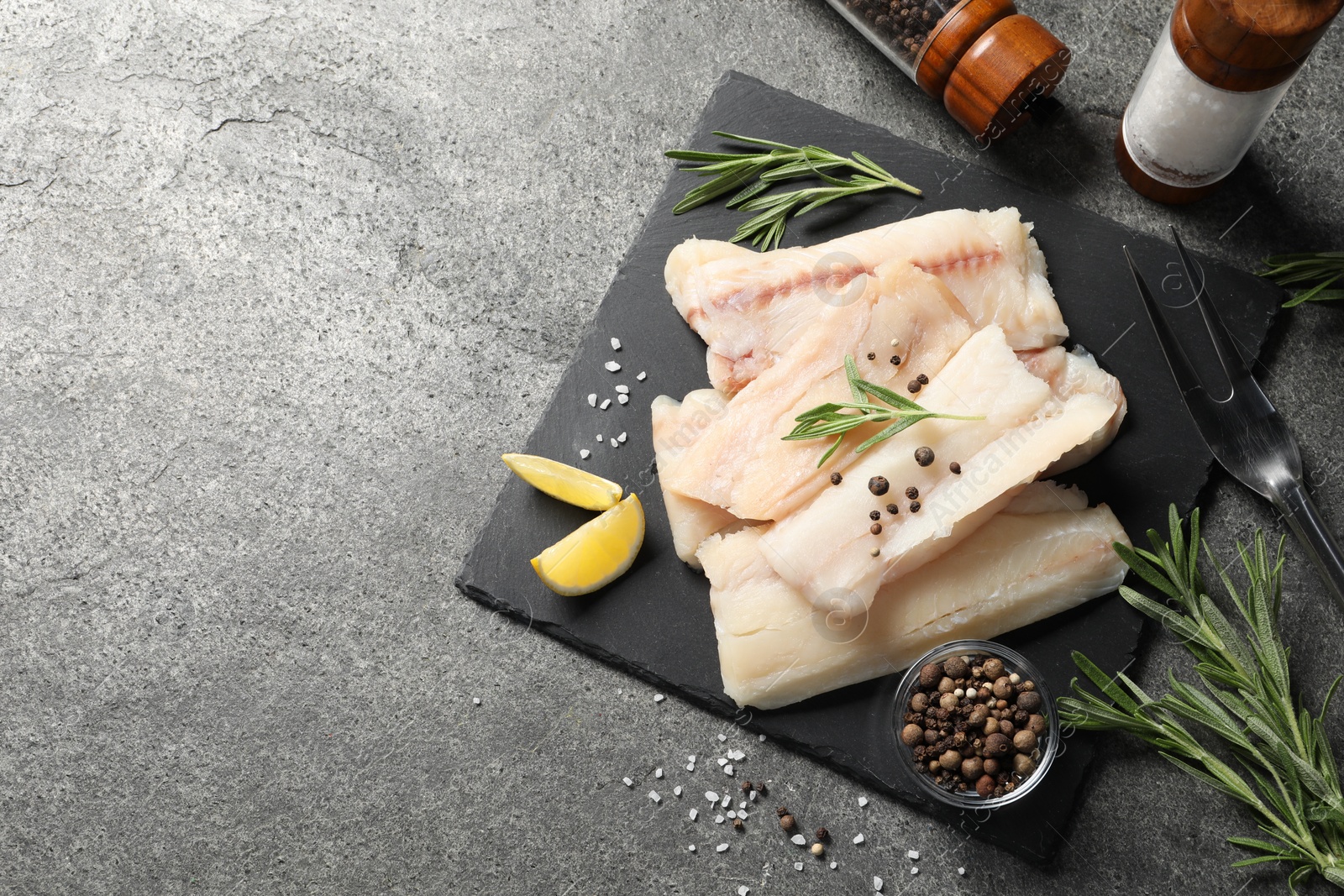 Photo of Pieces of raw cod fish, rosemary, peppercorns and lemon on grey table, top view. Space for text