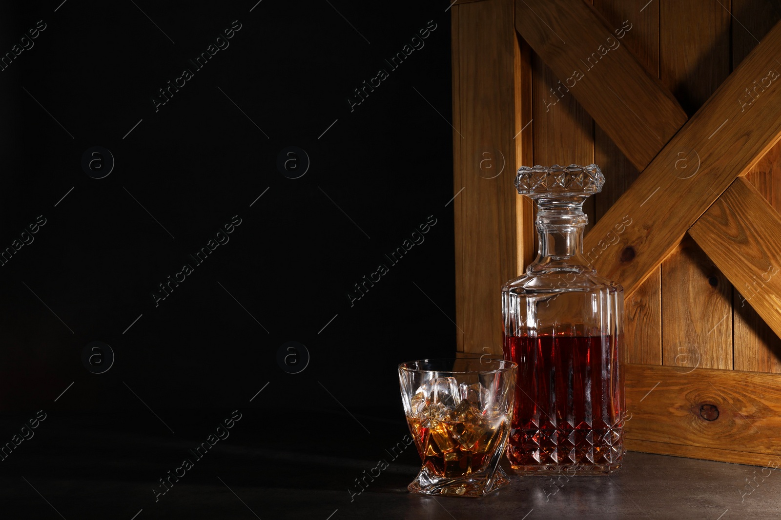 Photo of Whiskey in glass and bottle near wooden crate on dark table against black background. Space for text