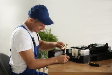 Photo of Repairman with magnifying glass and screwdriver fixing modern printer indoors