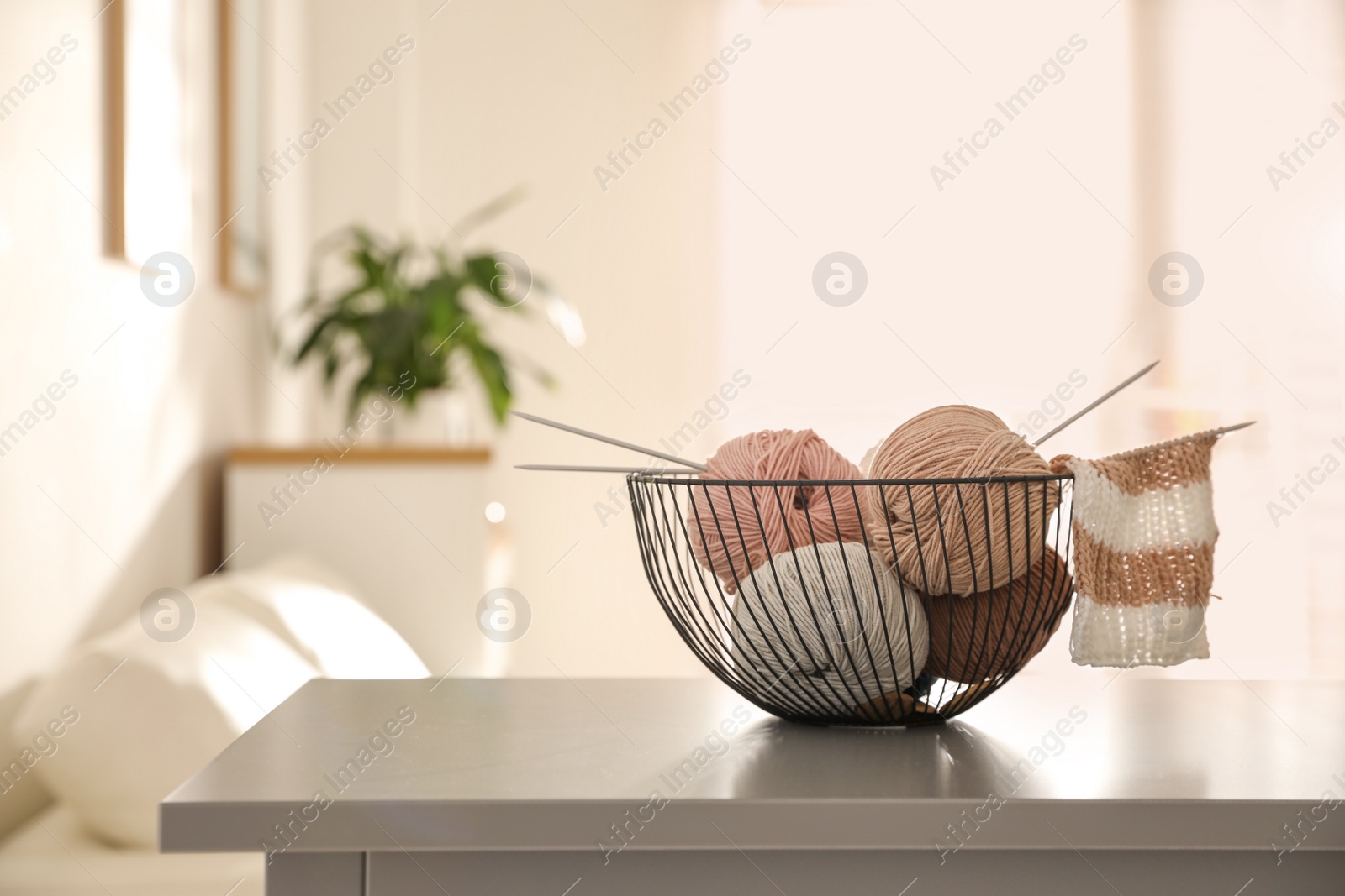 Photo of Yarn balls and knitting needles in metal basket on grey table indoors, space for text. Creative hobby