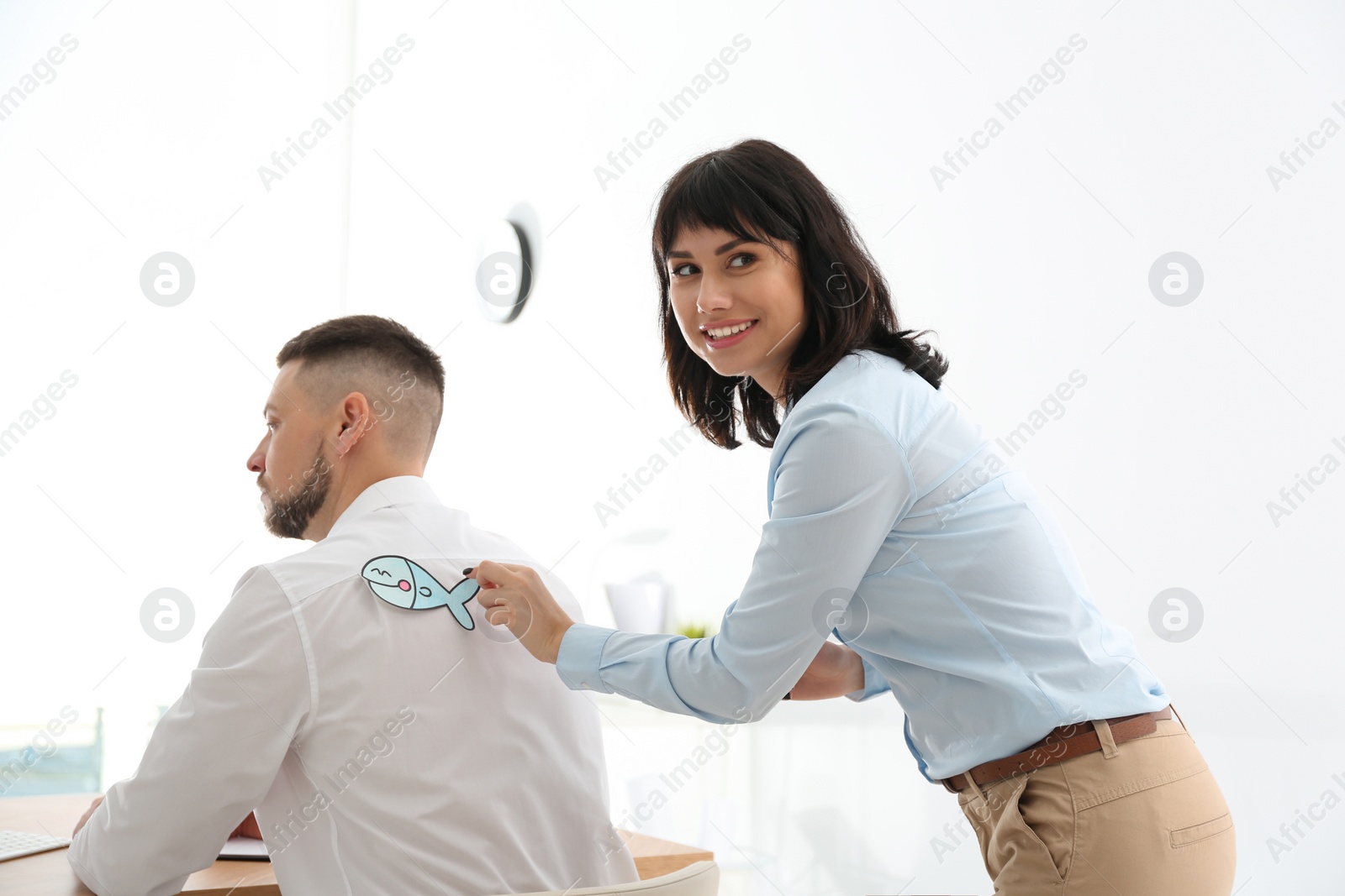 Photo of Young woman sticking paper fish to colleague's back in office. April fool's day