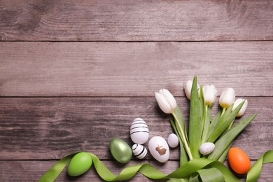 Many painted Easter eggs, tulip flowers and ribbon on wooden table, flat lay. Space for text