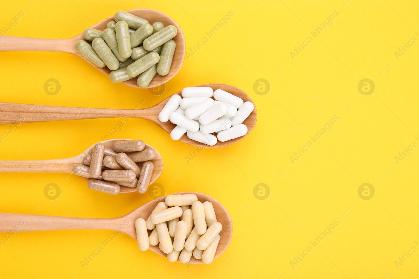 Photo of Different vitamin capsules in wooden spoons on yellow background, flat lay. Space for text