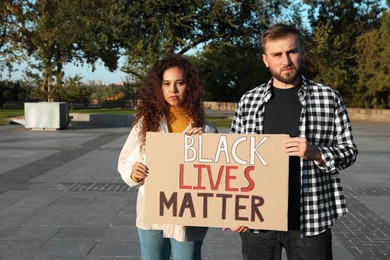 Photo of Young man and African American woman holding sign with phrase Black Lives Matter outdoors. Racism concept