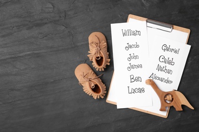 Clipboard with list of baby names, child's shoes and wooden toy on black table, flat lay. Space for text