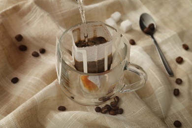 Photo of Pouring hot water into glass cup with drip coffee bag on beige fabric, closeup