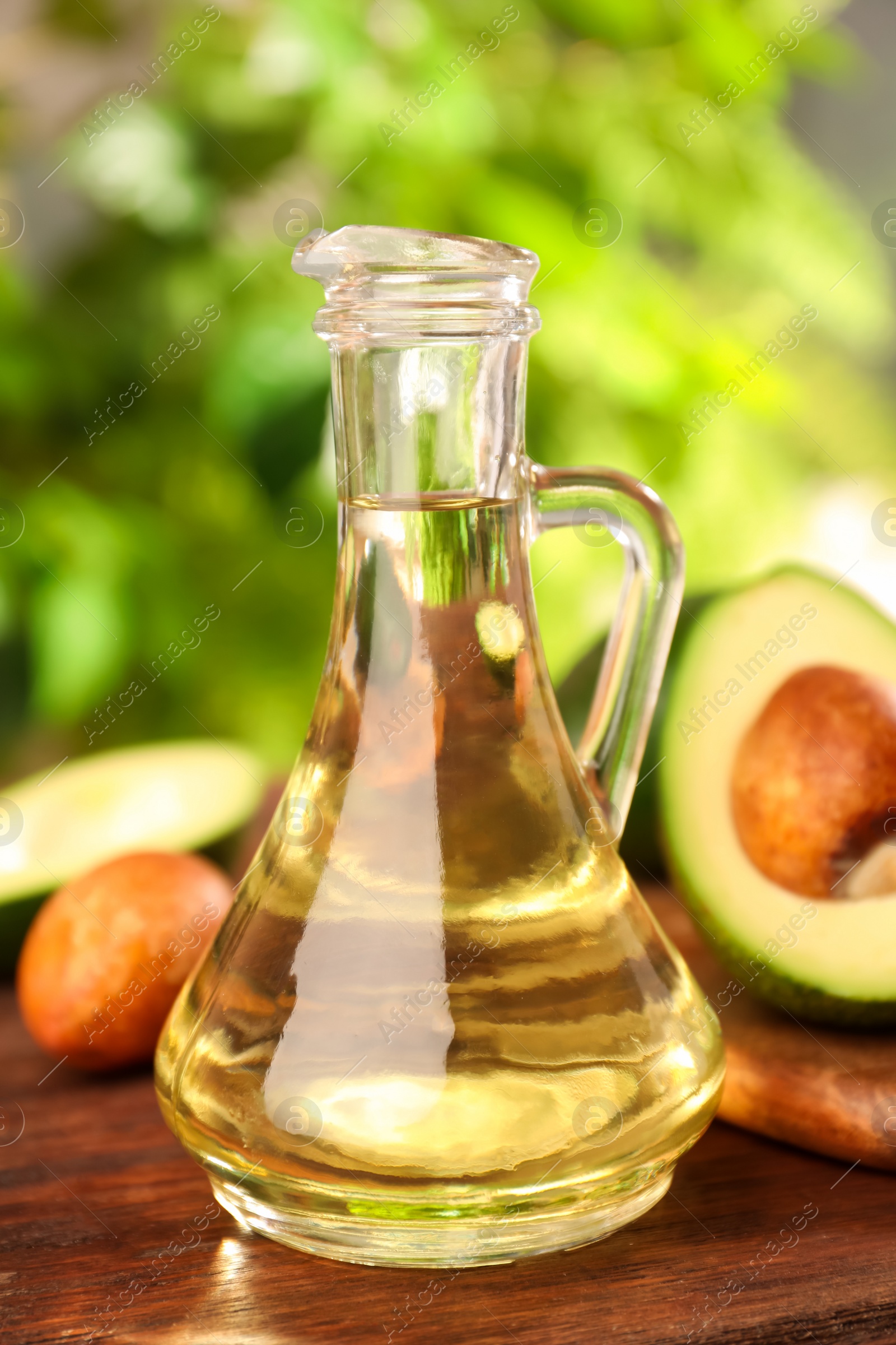 Photo of Glass jug of cooking oil and fresh avocados on wooden table against blurred green background