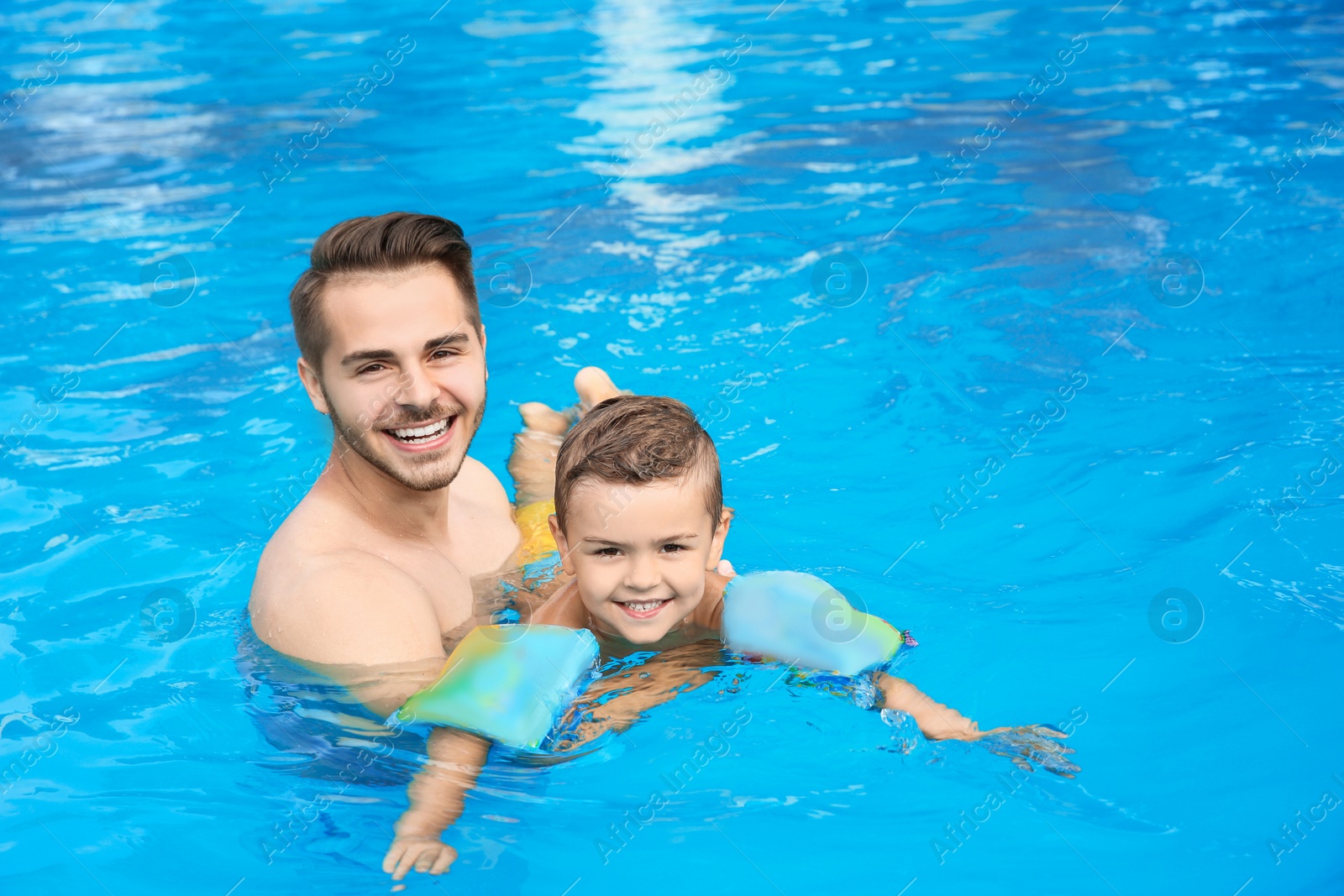 Photo of Father teaching son to swim with inflatable sleeves in pool on sunny day