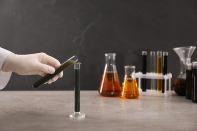 Photo of Woman pouring green crude oil into test tube at grey table, closeup