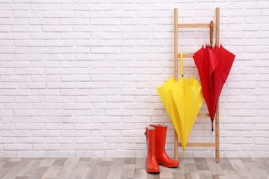 Photo of Stylish bright umbrellas on ladder and rubber boots near white brick wall. Space for text