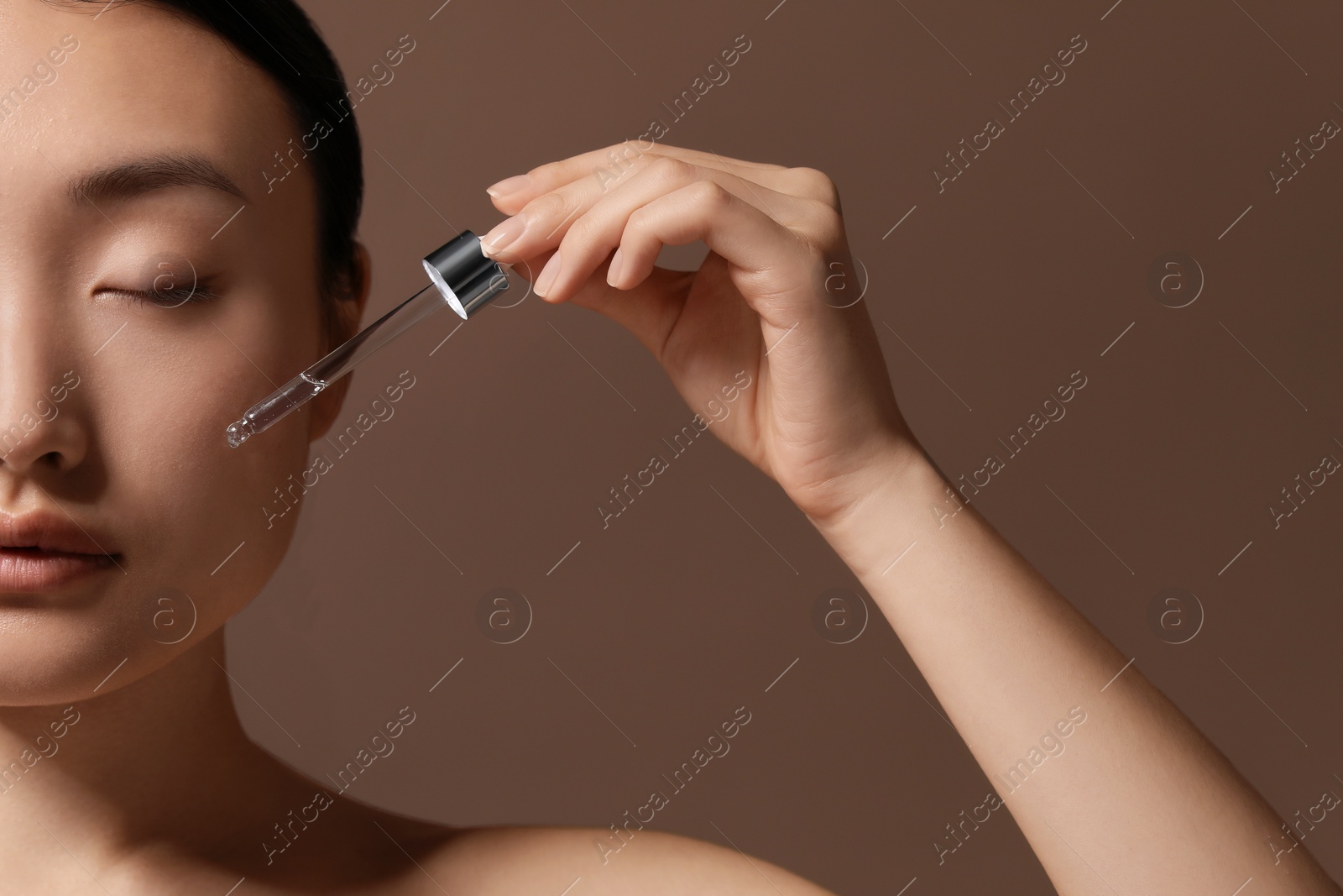 Photo of Beautiful young woman applying cosmetic serum onto her face on brown background, closeup