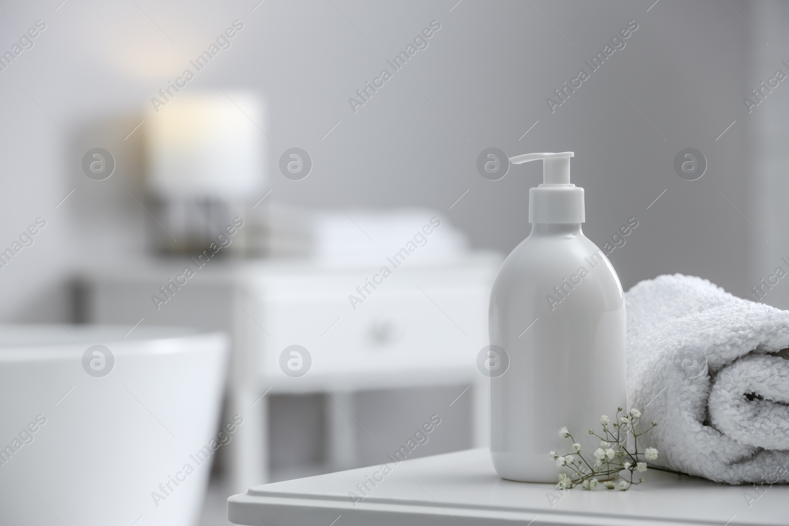 Photo of Bottle of bubble bath, towel and flowers on white table in bathroom, space for text