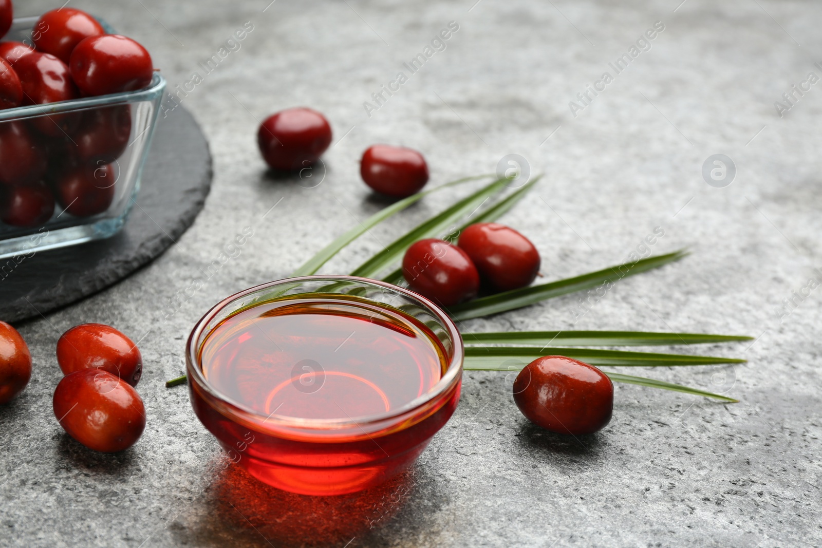 Photo of Palm oil in glass bowl, tropical leaf and fruits on grey table