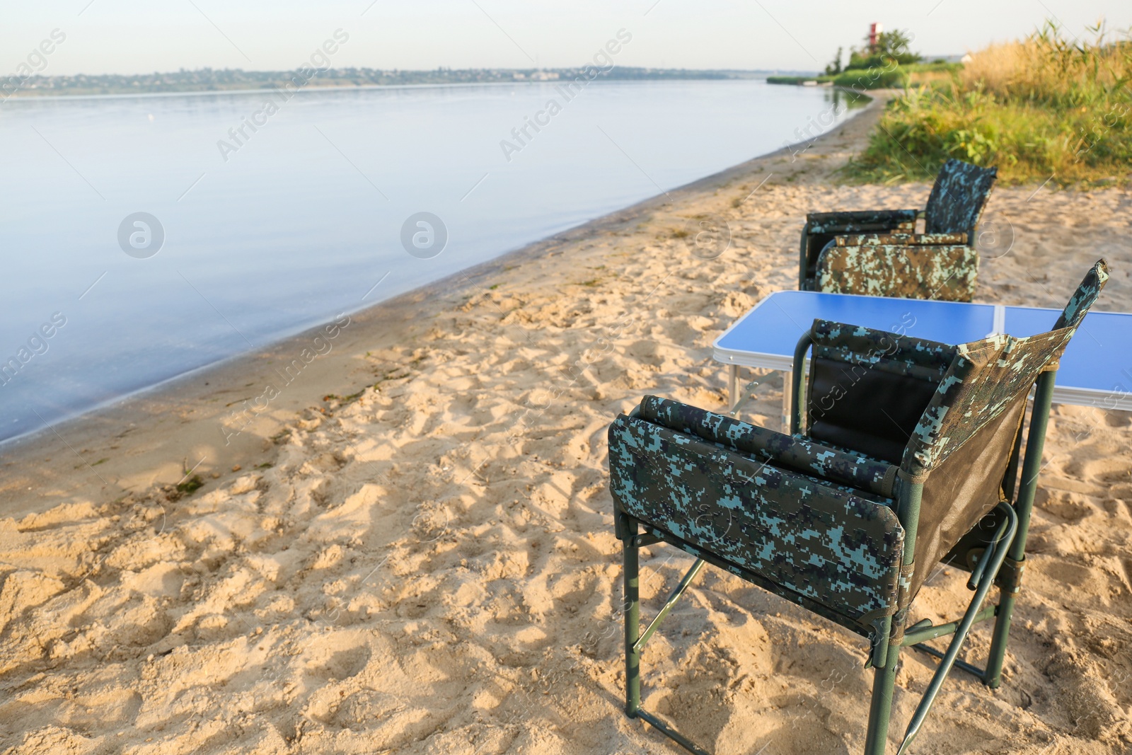 Photo of Camouflage fishing chairs and table on sandy beach near river, space for text