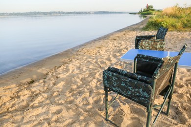 Photo of Camouflage fishing chairs and table on sandy beach near river, space for text