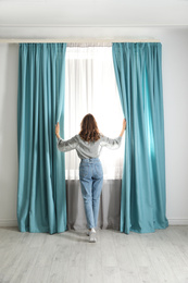 Photo of Woman opening window curtains at home in morning, back view