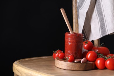 Photo of Jar of tasty tomato paste with spoon and ingredients on wooden table against black background, space for text