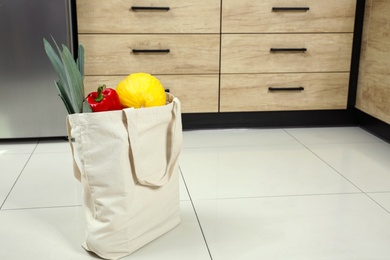 Photo of Tote bag with vegetables and fruits on floor in kitchen. Space for text