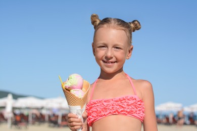 Photo of Adorable little girl in swimsuit with delicious ice cream at beach on sunny summer day