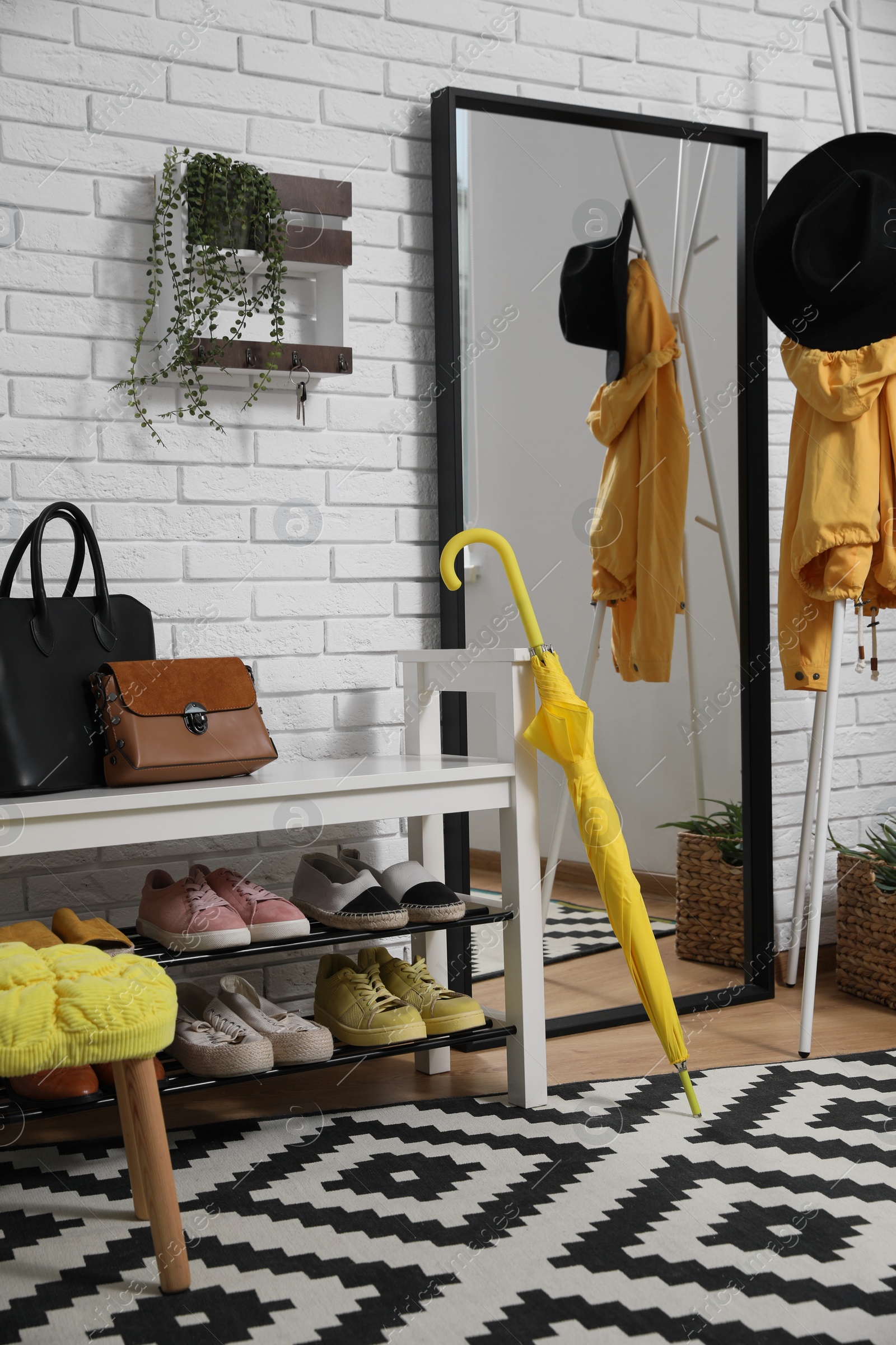 Photo of Stylish hallway interior with coat rack, shoe storage bench and mirror near white brick wall