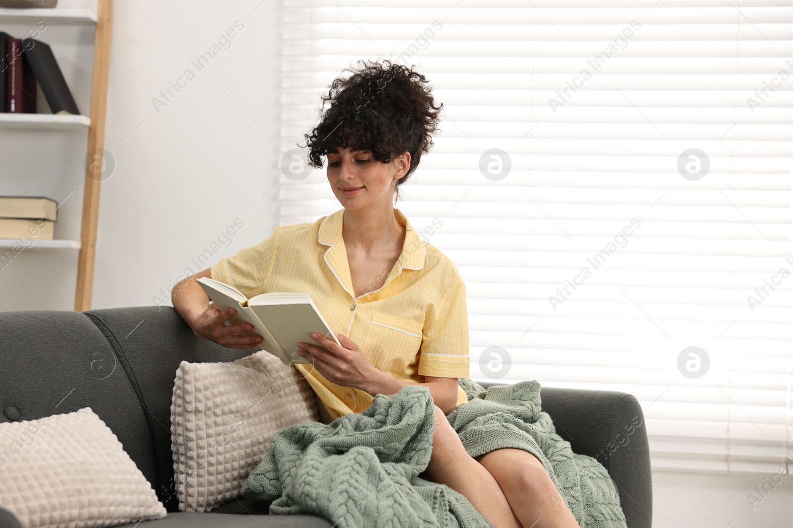 Photo of Beautiful young woman in stylish pyjama reading book on sofa at home