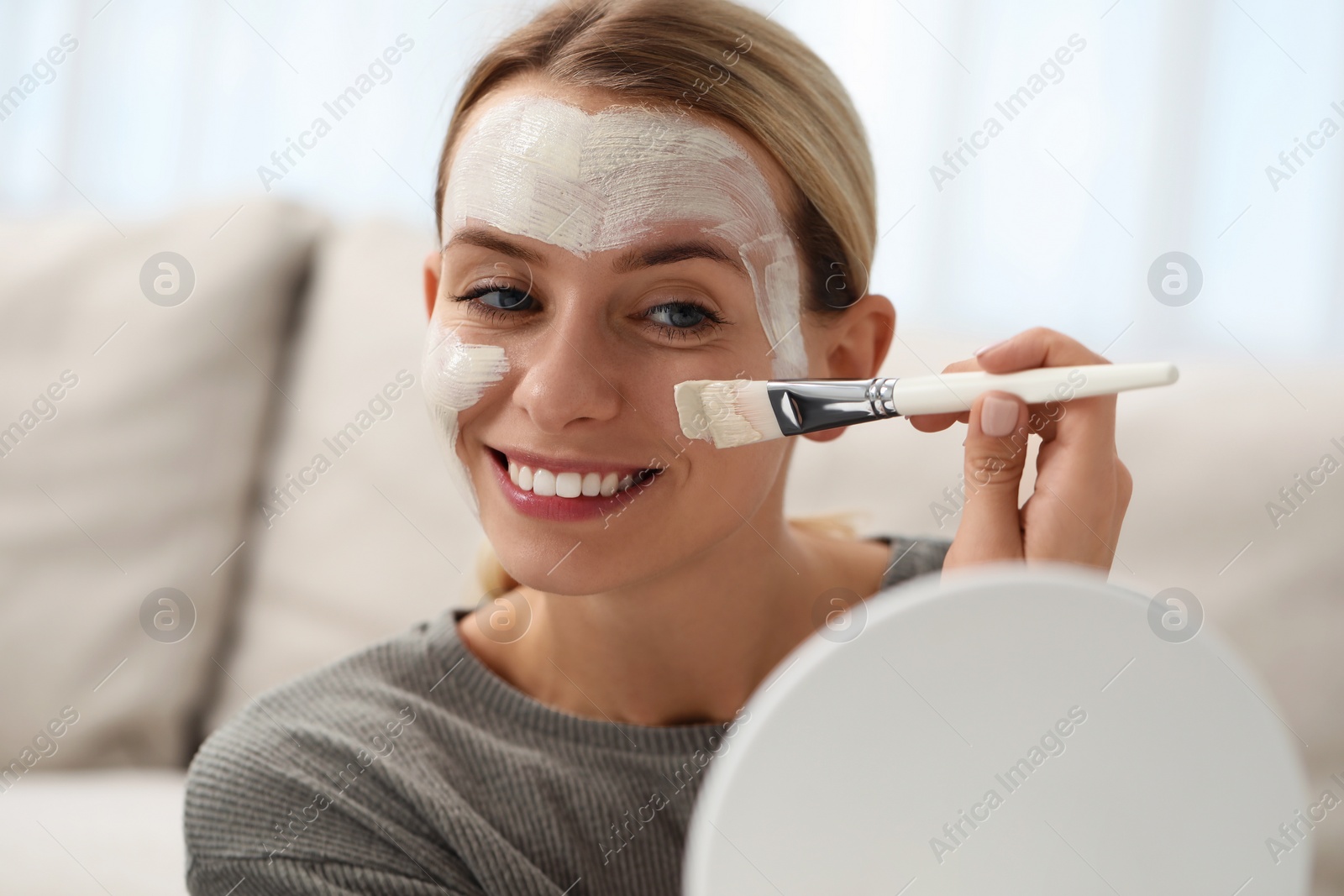 Photo of Young woman applying face mask in front of mirror indoors. Spa treatments