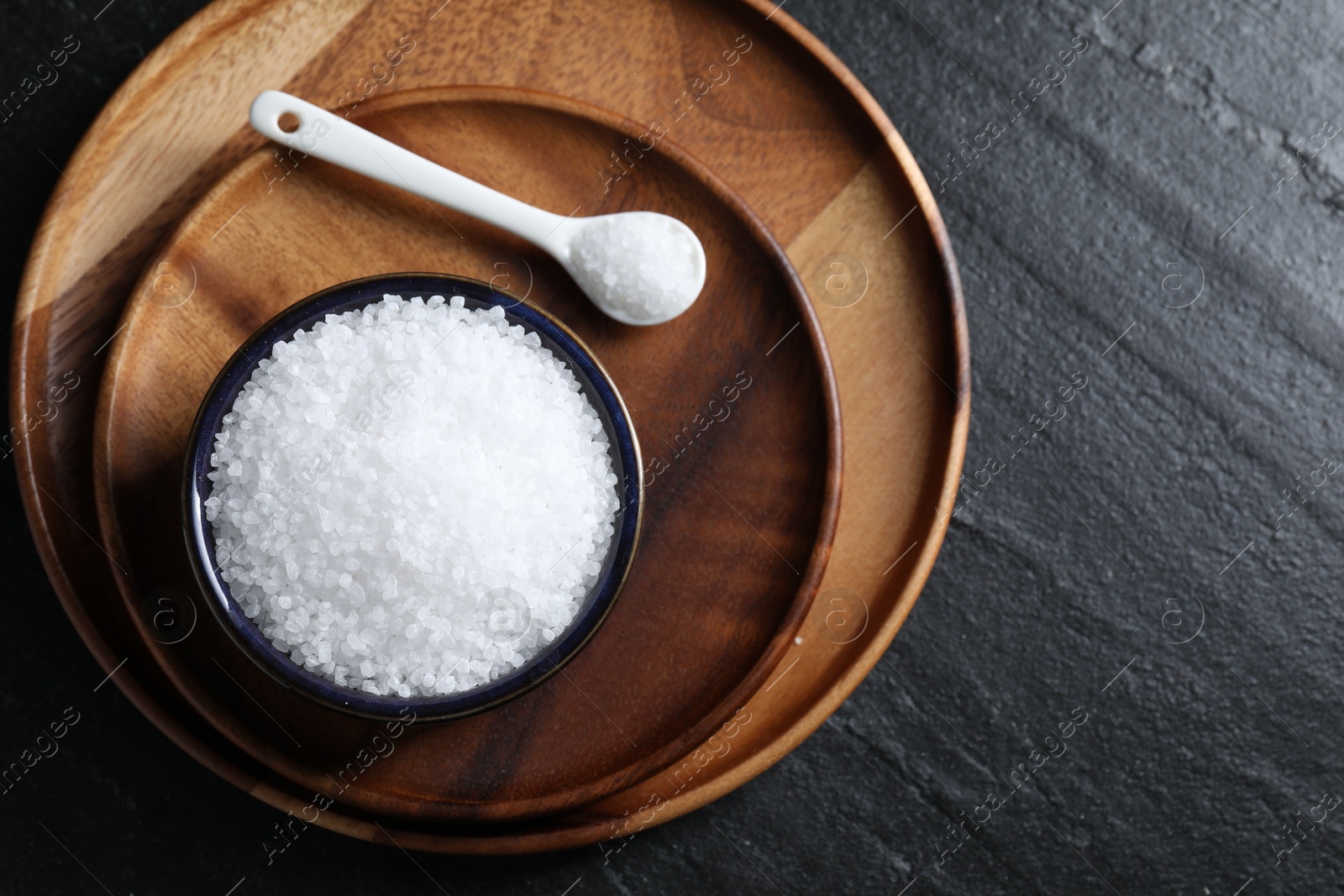 Photo of Organic white salt in bowl and spoon on black table, top view. Space for text