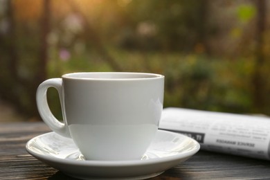 Photo of White cup with coffee and newspaper on wooden table, closeup. Morning ritual